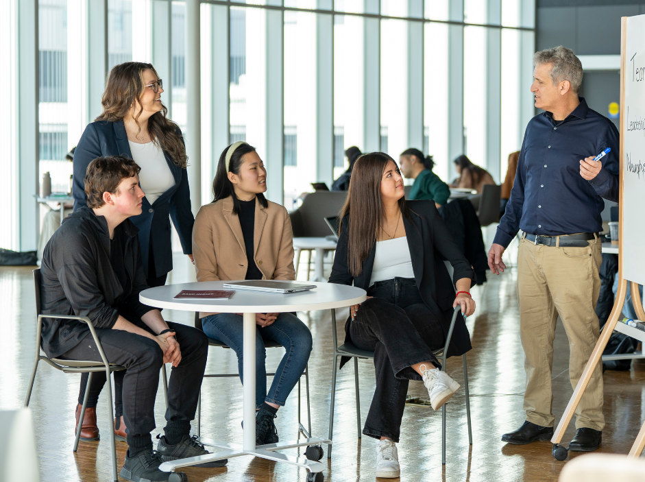 A small group people around a table paying attention to an instructor or leader at a whiteboard.